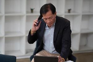 Asian senior manager and businessman in black suit relaxing in social media online and talking, calling with businesspeople on smartphone at workspace in office photo