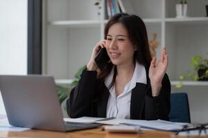 Young accountant business asian woman with smartphone to call marketing consultants and using laptop computer to analyze sales growth in the global workplace market. accounting concept. photo