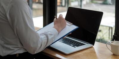 Business woman hand are taking notes on paper with a pen, and she is using a laptop computer on a wooden desk in the office.Web banner photo