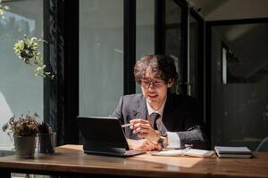 Asian young businessman in eyewear working with digital tablet remotely, sitting at wooden table in cafe. Pleasant happy man communicating in social network, searching information online photo