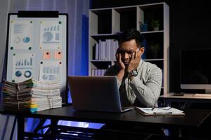 Tired or bored asian male worker sit at desk need rest suffer from fatigue overworked at office. Exhausted man working at computer late at night try to meet deadline on project photo