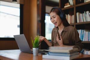 Asian tutor of language start online lesson with trainee wave hand smiling looks at pc screen. woman in headphones communicating distantly, e-learning process, application usage concept photo