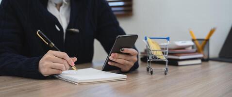 woman write in notebook use cellphone study online at home. Happy ethnic female take notes handwrite on paper with application on smartphone. Planning concept. photo