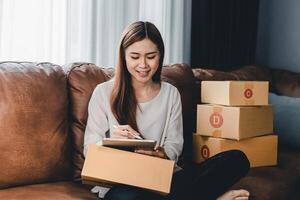 Asian female with small stock business owner holding digital tablet and retail package parcel boxes checking commercial shipping delivery order on smartphone using mobile app technology. photo