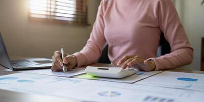 Close up of businesswoman or accountant hand holding pen working on calculator to calculate business data, accountancy document and laptop computer at office, business concept photo