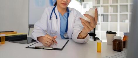 Female doctor writing in a notebook and holding a medicine bottle works at a computer while there were many medicine bottles on the table while sitting at a work desk photo