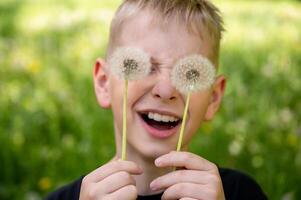 The boy has covered his eyes with dandelions and is smiling widely against a green background. Selective focus. photo
