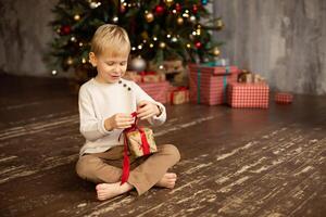 Cute little boy sitting on the floor in front of the Christmas tree with gifts photo