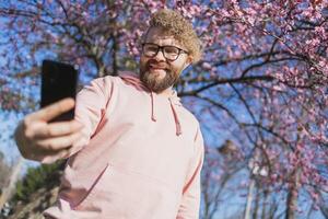 Happy curly man takes selfie against backdrop of flowering tree in spring for his internet communications. Weekend and social networks concept photo