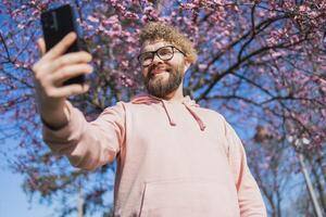 Happy curly man takes selfie against backdrop of flowering tree in spring for his internet communications. Weekend and social networks concept photo