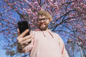 Happy curly man takes selfie against backdrop of flowering tree in spring for his internet communications. Weekend and social networks concept photo