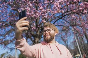 Happy curly man takes selfie against backdrop of flowering tree in spring for his internet communications. Weekend and social networks concept photo