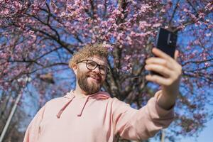 Happy curly man takes selfie against backdrop of flowering tree in spring for his internet communications. Weekend and social networks concept photo