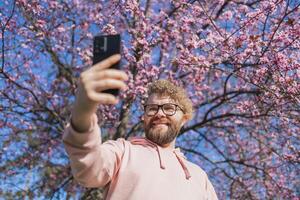 Happy curly man takes selfie against backdrop of flowering tree in spring for his internet communications. Weekend and social networks concept photo