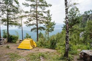 A yellow tent stands on the edge of a cliff in the mountains, a house for sleeping in nature, camping in the forest, a camp site, a tourist parking lot. photo