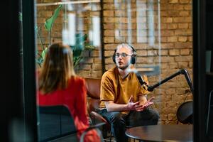 A gathering of young business professionals, some seated in a glass-walled office, engage in a lively conversation and record an online podcast, embodying modern collaboration and dynamic interaction photo