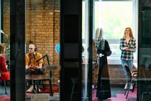 A gathering of young business professionals, some seated in a glass-walled office, engage in a lively conversation and record an online podcast, embodying modern collaboration and dynamic interaction photo