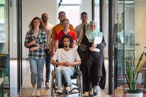 A diverse group of young business people congregates within a modern startup's glass-enclosed office, featuring inclusivity with a person in a wheelchair, an African American young man, and a hijab photo
