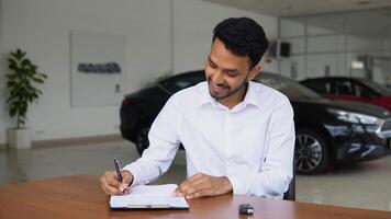 Indian salesman making deal signs a sales document of a car while sitting at table in dealership office video