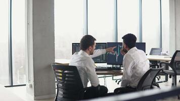 Two multiracial investors brokers analysing indexes online cryptocurrency stock. Diverse managers traders analysts discussing financial growth market at desk with pc graphs on screen video