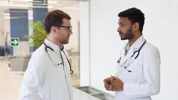 Medical multiethnic staff having discussion in a hospital hallway. Indian and caucasian doctors in white coat working in a medical clinic. Two hospital workers discussing on laboratory test video