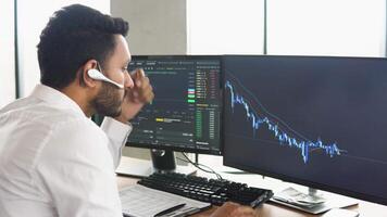 Side view of smart Indian crypto trader, investor, analyst broker, using pc analyzing digital cryptocurrency exchange, stock market charts, thinking of investing and funds risks video