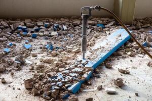 A pneumatic jackhammer demolishes walls and floor, creating a cloud of dust and debris. The powerful tool showcases intense construction work in progress. photo