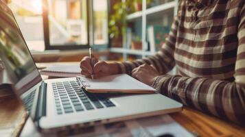 Person taking notes on a notepad while working on a laptop in a cozy, sunlit room, enhancing productivity and focus. photo