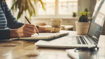 Person writing in a notebook near a laptop in a cozy workspace, sunlight filtering through the window creating a warm and productive atmosphere photo
