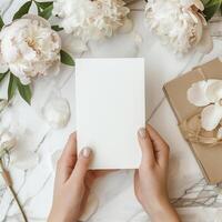 A woman's hands holding a blank notecard with a marble background and white and cream colored flowers and a present with a bow on the left side of the image. photo
