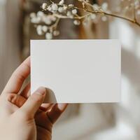 Minimalistic mockup of blank paper card holden by hand with blurred flowers in the background. photo