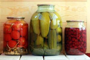 Three jars of pickled cucumbers, tomatoes and cherry jam against the wooden background photo
