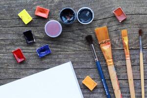 Flat layout of colorful acrylic and watercolor paints, set of brushes and bound white album sheet mock uo on rough grey wooden table texture photo