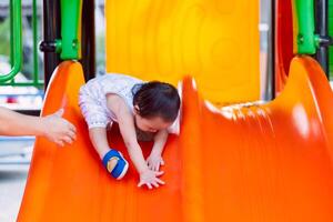 A little boy having fun on a playground slide during a sunny day. Cheerful little boy having fun while sliding outdoors. Parent's hands protecting little child. photo