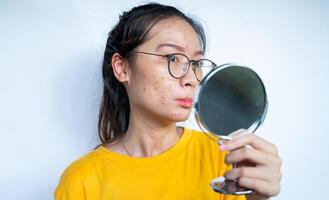 Young Asian woman feeling shock while looking acne and scar occur on face by mini mirror. Conceptual shot of Acne and Problem Skin on female face. photo