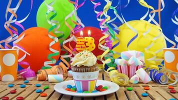 Birthday 83 cupcake with candles burning on rustic wooden table with background of colorful balloons, plastic cups and candies with blue wall in the background photo