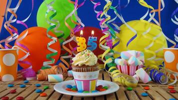 Birthday 35 cupcake with candles burning on rustic wooden table with background of colorful balloons, plastic cups and candies with blue wall in the background photo