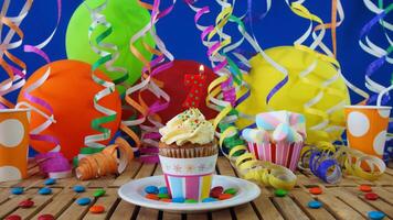Birthday 7 cupcake with candles burning on rustic wooden table with background of colorful balloons, plastic cups and candies with blue wall in the background photo