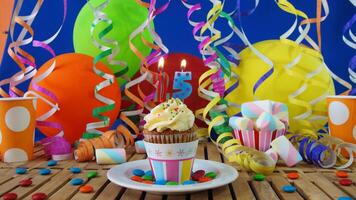 Birthday 25 cupcake with candles burning on rustic wooden table with background of colorful balloons, plastic cups and candies with blue wall in the background photo