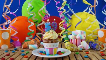 Birthday 59 cupcake with candles burning on rustic wooden table with background of colorful balloons, plastic cups and candies with blue wall in the background photo