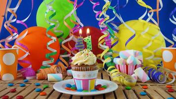 Birthday 41 cupcake with candles burning on rustic wooden table with background of colorful balloons, plastic cups and candies with blue wall in the background photo