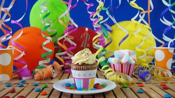 Birthday 2 cupcake with candles burning on rustic wooden table with background of colorful balloons, plastic cups and candies with blue wall in the background photo