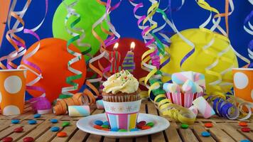 Birthday 24 cupcake with candles burning on rustic wooden table with background of colorful balloons, plastic cups and candies with blue wall in the background photo