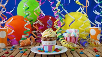 Birthday 9 cupcake with candles burning on rustic wooden table with background of colorful balloons, plastic cups and candies with blue wall in the background photo