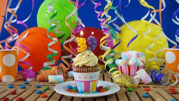 Birthday 39 cupcake with candles burning on rustic wooden table with background of colorful balloons, plastic cups and candies with blue wall in the background photo