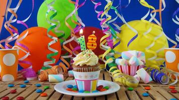 Birthday 18 cupcake with candles burning on rustic wooden table with background of colorful balloons, plastic cups and candies with blue wall in the background photo