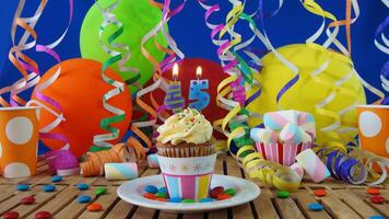 Birthday 45 cupcake with candles burning on rustic wooden table with background of colorful balloons, plastic cups and candies with blue wall in the background photo
