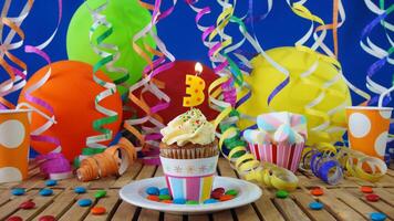 Birthday 3 cupcake with candles burning on rustic wooden table with background of colorful balloons, plastic cups and candies with blue wall in the background photo