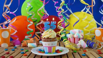 Birthday 55 cupcake with candles burning on rustic wooden table with background of colorful balloons, plastic cups and candies with blue wall in the background photo