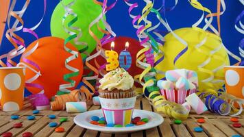 Birthday 30 cupcake with candles burning on rustic wooden table with background of colorful balloons, plastic cups and candies with blue wall in the background photo